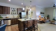 This single-story space showcases an open-concept layout that integrates the kitchen and living area into one cohesive, functional environment. The kitchen is a standout feature, centered around a granite-topped island with seating for two, accented by stylish pendant lighting that hangs overhead. Rich, dark wood cabinetry lines the walls, complementing the warm hardwood flooring that extends throughout the kitchen and transitional spaces. Stainless steel appliances, including a full-sized refrigerator, add a modern touch, while the sink area is framed by ample counter space and additional upper cabinetry for storage. Built-in open shelving provides a decorative display area, adding visual interest to the layout. The transition from kitchen to living area is seamless, with soft carpeting defining the lounge space, where warm ambient lighting from a table lamp creates an inviting atmosphere. Recessed lighting and crown molding details throughout contribute to the polished, well-finished feel of the interior. The overall ambiance is warm and welcoming, combining practical design with tasteful aesthetic choices.