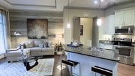 This single-floor residence showcases a thoughtfully designed open-concept layout that seamlessly blends the kitchen and living areas into one cohesive, inviting space. The kitchen is a standout feature, boasting dark granite countertops that extend into a generous breakfast bar, complete with sleek modern bar stools and elegant pendant lighting that casts a warm, focused glow. White cabinetry and a classic farmhouse-style sink complete the polished culinary space.

The adjoining living area is anchored by rich hardwood flooring and a striking accent wall clad in weathered wood paneling, adding warmth and texture to the space. A large-scale abstract artwork mounted above the sofa serves as a sophisticated focal point, complementing the neutral, contemporary color palette. The living room is furnished with a plush light-gray sofa, a patterned area rug, and a glass coffee table, creating a comfortable and stylish gathering area.

Recessed tray ceiling details add architectural depth, while the open sightlines between rooms enhance the sense of space and airflow. The overall ambiance is modern yet warm, blending contemporary design elements with inviting textures and curated décor for a refined, livable aesthetic.