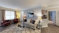 This single-floor residence offers a thoughtfully designed layout with distinct living spaces that flow naturally into one another. The kitchen is outfitted with dark espresso cabinetry, granite-look countertops, stainless steel appliances including a French door refrigerator, and wood-look flooring that extends into the main living areas. A dedicated dining area features crown molding and a pendant light fixture overhead, creating an inviting space for meals and gatherings. The living room is generously sized, featuring crown molding, wood-look flooring, and a built-in media niche with cabinet storage below — a practical and stylish focal point. Natural light filters in through windows dressed with decorative curtains, lending warmth to the neutral color palette throughout. Two bedrooms provide comfortable retreats; one features a tray ceiling with crown molding detail and plush carpeting, while both rooms offer ample space and access to storage. A large walk-in closet with wire shelving and hanging rods provides excellent organization capacity. The two bathrooms are well-equipped — one includes a tub/shower combination with classic tile accents, and the other offers a wide vanity with a large mirror and bright overhead lighting. Overall, the space presents a clean, modern aesthetic with cohesive finishes throughout.