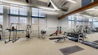 Located at 57 Charter Oak Ave in Hartford, Connecticut, this fitness facility offers a spacious and well-appointed workout environment spread across a single floor. The space comprises two exercise rooms and connecting hallways, providing ample room for a full range of training activities.

The exercise rooms showcase an industrial-inspired aesthetic, featuring soaring ceilings with exposed silver ductwork, rich wood beam accents, and floor-to-ceiling multi-pane windows that flood the space with abundant natural light. Neutral-toned carpeting runs throughout, complementing the white brick and two-tone painted walls.

The primary exercise room is generously sized and lined with floor-to-ceiling mirrors, creating an open and expansive feel. It is outfitted with a wide selection of strength-training equipment, including cable machines, weight benches, a dumbbell rack, medicine ball stations, and rowing machines. The second exercise room continues the same design language and houses an array of selectorized strength machines and cardio equipment, including stationary bikes and treadmills. Wall-mounted televisions and decorative artwork add a polished finish to the space.

Linear LED lighting runs along the ceilings, ensuring the entire facility is brightly and evenly lit. The overall ambiance is clean, modern, and functional, making this a comprehensive fitness destination in the heart of Hartford.