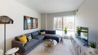 Located on the 8th floor at 710 12th St in Arlington, Virginia, this well-appointed unit offers a thoughtfully designed single-floor layout with modern finishes and sweeping city views. The entry foyer features a rich wood-toned door with a sleek lever handle, setting a contemporary tone from the moment you arrive. Light hardwood flooring runs through the main living areas, complementing the clean, neutral palette throughout.

The open kitchen is a standout feature, showcasing two-tone cabinetry — dark walnut lowers paired with crisp white uppers — alongside stainless steel appliances, a tile backsplash, and a generous island with pendant lighting and bar seating. The kitchen flows seamlessly into the dining and living areas, making it ideal for entertaining.

The living room is bathed in natural light from floor-to-ceiling windows that frame expansive views of the Arlington skyline. A large sectional sofa and bold abstract artwork add character to the bright, airy space.

The bedroom continues the theme of comfort and style, featuring large windows with city views, soft neutral tones, a statement brass chandelier, and warm mid-century modern furnishings. The bathroom offers a clean, contemporary design with navy cabinetry and light tile flooring. Every detail of this space reflects a cohesive, modern aesthetic.