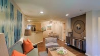 This single-floor residence offers a thoughtfully designed open-concept layout that effortlessly connects the living, dining, and kitchen areas into one cohesive and inviting space. The living area is warm and welcoming, featuring rich hardwood-style flooring, recessed lighting, and a ceiling fan that adds both style and functionality. A decorative accent wall with a round statement mirror anchors one side of the room, complemented by a rustic media console that adds character and warmth. The space comfortably accommodates a plush seating arrangement accented with vibrant orange throw pillows, centered around a woven coffee table that brings a natural, textural element to the room. Just beyond, a compact dining nook features a round table and modern chairs beneath soft ambient light, creating an intimate setting for meals. The kitchen is open to the living space via a breakfast bar counter, offering casual seating and a sense of connectivity. It is equipped with stainless steel appliances and light wood cabinetry, providing a clean and functional cooking environment. Built-in shelving near the dining area adds convenient display or storage space. The overall ambiance is bright, airy, and cohesive, with neutral tones throughout and carefully chosen décor that creates a comfortable and stylish atmosphere.