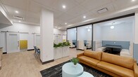 Located at 237 William Howard Taft Rd in Cincinnati's Uptown neighborhood, this thoughtfully designed coworking space offers a well-rounded environment for productivity and collaboration across a single floor. The open-plan layout is anchored by a central lounge area featuring a warm cognac leather sofa, light blue accent tables, and lush greenery, creating an inviting focal point within the space. Light wood flooring runs throughout, complemented by a drop-tile ceiling with recessed lighting that keeps the atmosphere bright and professional. A row of semi-private glass-enclosed booths along one side provides seating nooks ideal for small meetings or focused work, each furnished with upholstered seating and pendant lighting. Multiple private workrooms feature a distinctive two-tone design — bold mustard yellow lower walls paired with neutral upper walls — along with a clean white desk, ergonomic chair, and a blue gooseneck wall sconce. A collaborative meeting room includes a large wood table with blue chairs, built-in cabinetry, and a printer station set against a muted blue accent wall. Sliding barn-style doors, structural white columns, and built-in bench seating along the perimeter complete the space, which also includes clearly marked safety and exit infrastructure for occupant convenience.
