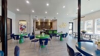 This single-story community clubroom offers a generously sized, open-plan gathering space thoughtfully designed for both socializing and relaxation. The layout is anchored by a striking lounge area furnished with vibrant green and blue round tables paired with quilted navy chairs, creating an energetic yet cohesive color palette throughout. Large-format light tile flooring runs continuously across the space, enhancing its open, airy feel.

A standout feature is the dark tile accent wall housing a wall-mounted television above a sleek linear gas fireplace, flanked by warm pendant sconces that add an inviting ambiance. Adjacent to the lounge, a fully equipped kitchenette serves as a functional hub, featuring granite-topped counters with tiled bases, white cabinetry, a stainless steel range, and a commercial-grade refrigerator — ideal for catered events or community gatherings.

A glass-walled private meeting or dining room sits along one side, flooded with natural light through a series of large windows overlooking an outdoor pool and courtyard. Bold artwork adorns the walls, complementing the contemporary interior design. Recessed lighting throughout ensures the space remains bright and welcoming at any time of day.