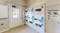 Located at 400 Greenlawn Dr in Columbia, South Carolina, this single-floor shared laundry facility is designed to serve the needs of its users efficiently and conveniently. The room is laid out to maximize functionality, with a row of top-loading washing machines lining one wall, each equipped with individual digital payment terminals for easy, cashless operation. Opposite the washers, stacked dryer units provide ample drying capacity, also outfitted with card-based payment systems. Natural light filters into the space through a window fitted with horizontal blinds, creating a bright and welcoming environment. The room features durable, easy-to-clean speckled flooring and neutral-toned walls that contribute to a clean, utilitarian atmosphere. A fold-down table with a chair offers a dedicated folding and waiting area, making the laundry experience more comfortable. Informational signage is posted throughout, including instructions for using a mobile app to manage laundry cycles remotely. An electrical panel is mounted on the wall near the exit door, which provides direct access to the surrounding outdoor area. Overall, the facility is well-organized, practical, and equipped with modern amenities to streamline the laundry process.