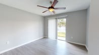 This single-floor unit is finished with a consistent, clean aesthetic throughout. Light hardwood-style flooring runs continuously across all rooms, complemented by crisp white baseboards and neutral gray walls that create a bright, cohesive atmosphere. Textured ceilings add subtle visual depth to the living spaces.

The living area is open and well-proportioned, connecting directly to the kitchen via a pass-through counter with overhead cabinetry finished in warm tones. This arrangement allows for easy interaction between the two spaces while maintaining a defined layout. The kitchen includes light-colored cabinetry and built-in appliances.

Multiple rooms are equipped with ceiling fans, providing both airflow and ambient lighting. A standout feature of the unit is the sliding glass door access point, framed by vertical blinds, which opens to an outdoor area and offers a picturesque view of a water feature and surrounding greenery, flooding the interior with natural light.

The entry area includes a closet alongside the front door, offering convenient storage. The bathroom is neatly appointed with a white vanity featuring a speckled countertop, a tub-shower combination with tiled surround, and matching hardwood-style flooring that ties the space together with the rest of the unit.