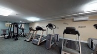 This fitness center offers a comprehensive workout environment designed to accommodate a wide range of exercise needs. The space is generously proportioned, with a long, open floor plan that allows for comfortable movement between equipment zones. Durable, multi-tonal carpet flooring covers the entire area, providing both practicality and cushioning underfoot.

The cardio section is prominently featured and includes multiple Matrix treadmills, elliptical trainers, stair climbers, and stationary bikes — including a recumbent model — lined along the walls to maximize open floor space. A dedicated strength training area houses a multi-station cable machine and a flat weight bench, catering to resistance-based workouts.

A functional training corner adds versatility to the space, stocked with an assortment of stability balls, medicine balls, resistance bands, a BOSU balance trainer, and foam rollers organized neatly on racks. A wall-mounted flat-screen TV is positioned for convenient viewing during workouts.

The walls are adorned with motivational text decals reading "GYM," "FITNESS," "WIN," "SWEAT," and "CHALLENGE," reinforcing an energizing atmosphere. Overhead fluorescent and recessed lighting ensures the entire space is well-lit. A wall-mounted air conditioning unit supports climate control. The facility is accessed through a keyed entry door, adding a layer of security and exclusivity.