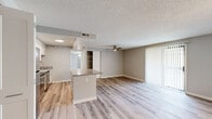 This single-story unit offers three bedrooms and one bathroom across a thoughtfully arranged floor plan. Light wood-style laminate flooring runs throughout, creating a cohesive, contemporary feel. The open-concept kitchen and living area forms the heart of the unit, where a peninsula with stone-look countertops separates the two spaces. The kitchen is equipped with stainless steel appliances—including a refrigerator, range, microwave, and dishwasher—alongside crisp white cabinetry and a stainless steel sink. The living room benefits from a ceiling fan and a sliding glass door fitted with vertical blinds, providing access to the outdoors and allowing natural light to fill the space. A built-in shelving unit adds functional storage near the kitchen passthrough. The three bedrooms feature neutral gray walls and light flooring; the generously sized primary bedroom includes a multi-bulb light fixture and ample wall space. The bathroom is finished with tile flooring, a white vanity with dark granite countertop, and a tiled tub-shower combination. Textured ceilings are present throughout most of the unit.