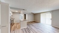 This single-story unit offers three bedrooms and one bathroom across a thoughtfully arranged floor plan. Light wood-style laminate flooring runs throughout, creating a cohesive, contemporary feel. The open-concept kitchen and living area forms the heart of the unit, where a peninsula with stone-look countertops separates the two spaces. The kitchen is equipped with stainless steel appliances—including a refrigerator, range, microwave, and dishwasher—alongside crisp white cabinetry and a stainless steel sink. The living room benefits from a ceiling fan and a sliding glass door fitted with vertical blinds, providing access to the outdoors and allowing natural light to fill the space. A built-in shelving unit adds functional storage near the kitchen passthrough. The three bedrooms feature neutral gray walls and light flooring; the generously sized primary bedroom includes a multi-bulb light fixture and ample wall space. The bathroom is finished with tile flooring, a white vanity with dark granite countertop, and a tiled tub-shower combination. Textured ceilings are present throughout most of the unit.