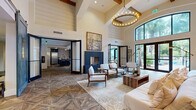 This thoughtfully designed amenity lounge spans a generous single floor, offering a sophisticated blend of modern and rustic aesthetics throughout its open, flowing layout. The main lounge area commands attention with its dramatic soaring ceilings, exposed wood beam accents, and a striking circular chandelier that anchors the space. A bold navy blue fireplace surround serves as a focal point, flanked by elegant white armchairs and a reclaimed wood coffee table set atop a textured area rug. Geometric parquet-style flooring in warm wood tones runs throughout, adding visual depth and continuity across each zone. Floor-to-ceiling black-framed windows and sliding glass doors flood the interior with natural light while offering sweeping views of lush outdoor greenery and a pool area. A dedicated lounge zone features plush white seating, open industrial shelving, and large-format wall art. The fully equipped kitchen showcases dark cabinetry, stainless steel appliances, a mosaic tile backsplash, a warm wood-paneled ceiling, and an island with bar seating. A cozy dining nook with a tufted banquette and pendant lighting completes the space, providing an inviting spot for casual gatherings.