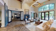 This thoughtfully designed amenity lounge spans a generous single floor, offering a sophisticated blend of modern and rustic aesthetics throughout its open, flowing layout. The main lounge area commands attention with its dramatic soaring ceilings, exposed wood beam accents, and a striking circular chandelier that anchors the space. A bold navy blue fireplace surround serves as a focal point, flanked by elegant white armchairs and a reclaimed wood coffee table set atop a textured area rug. Geometric parquet-style flooring in warm wood tones runs throughout, adding visual depth and continuity across each zone. Floor-to-ceiling black-framed windows and sliding glass doors flood the interior with natural light while offering sweeping views of lush outdoor greenery and a pool area. A dedicated lounge zone features plush white seating, open industrial shelving, and large-format wall art. The fully equipped kitchen showcases dark cabinetry, stainless steel appliances, a mosaic tile backsplash, a warm wood-paneled ceiling, and an island with bar seating. A cozy dining nook with a tufted banquette and pendant lighting completes the space, providing an inviting spot for casual gatherings.