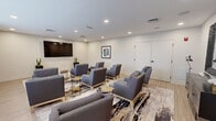This well-appointed lounge space offers a clean, contemporary design suited for group meetings, social gatherings, or media viewing. The room is finished with light hardwood flooring and crisp white walls, creating a bright and inviting atmosphere. Recessed lighting throughout the ceiling provides consistent, even illumination across the entire space.

The seating area is furnished with multiple coordinating gray upholstered armchairs arranged in a social configuration around a patterned area rug with abstract detailing. Slender-legged side tables in black and gold tones are distributed between the chairs, offering convenient surfaces for personal items. A large wall-mounted flat-screen television anchors one end of the room, making the space well-suited for presentations or media viewing.

A series of windows along one wall, fitted with light-filtering roller shades, allows diffused natural light to enter while maintaining privacy. Decorative framed artwork on the walls and potted plants in tall dark planters add refined finishing touches to the overall aesthetic. The layout is versatile, supporting both focused group interaction and relaxed informal gatherings. The space conveys a polished, professional ambiance with a comfortable residential quality.
