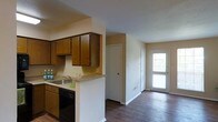 This single-floor space features a functional, open-concept kitchen that flows directly into the main living and hallway areas. The kitchen is equipped with warm-toned wooden cabinetry offering ample upper storage, complemented by a countertop with a built-in sink and a peninsula-style breakfast bar that visually separates the kitchen from the rest of the space. A black dishwasher is integrated beneath the counter, contributing to a practical layout. The countertops feature a speckled finish, and the backsplash area is finished in a neutral tone that keeps the space feeling clean and simple. Dark hardwood flooring runs throughout the hallway and adjoining areas, providing a grounding contrast to the light-colored walls. The hallway leads past an interior door, likely accessing a closet or utility space, toward a glass-paneled exterior door at the far end that allows natural light to filter into the space. The overall ambiance is straightforward and functional, with a neutral color palette and practical finishes throughout that make the space easy to personalize and maintain.