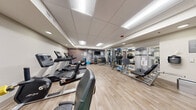 The Fitness Center at 500 Apex Dr in Windsor, Colorado, is a thoughtfully designed exercise facility that offers a comprehensive range of equipment within a single, efficiently organized room. The space features light hardwood-style flooring that contributes to a clean, modern aesthetic, while a drop-panel ceiling with recessed and panel lighting ensures the room is bright and well-illuminated throughout.

A standout design element is the bold, graphic accent wall featuring an abstract circular pattern in gray, white, and orange tones, lending the space a dynamic and energetic atmosphere. Cardio equipment lines one side of the room and includes multiple treadmills, an elliptical trainer, a recumbent bike, and a stair climber, all from Life Fitness. On the opposing side, a cable machine system and a multi-functional strength station accommodate a wide variety of resistance training exercises.

The free weights area is well-stocked, featuring a full rack of dumbbells, kettlebells of varying weights, and an assortment of medicine and stability balls. A wall-mounted flat-screen TV and posted workout guides add convenience for users. Floor-to-ceiling mirrors near the strength equipment enhance the sense of space and allow for proper form monitoring. The overall layout is practical and spacious, supporting both cardio and strength training needs.