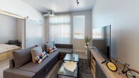 This single-floor unit features a contemporary design with a neutral gray-and-white color palette and light wood-style laminate flooring throughout. The kitchen is efficiently laid out, equipped with stainless steel appliances including a refrigerator, electric range, and over-the-range microwave. Gray cabinetry, open shelving, and a clean white countertop contribute to the kitchen's modern aesthetic, while an adjacent dining area provides a dedicated space for meals.

The main living area is generously sized, illuminated by natural light from large windows with horizontal blinds, and features track lighting overhead. A secondary, more intimate lounge space connects to the main area and includes wall-mounted climate control and additional window access.

The bedroom is a well-proportioned retreat with direct access to the en-suite bathroom. The bathroom includes a single-sink vanity with a white countertop, open shelving, a large mirror, and a glass-enclosed walk-in shower. Light-toned flooring carries consistently through all spaces, reinforcing a cohesive, clean look. Overall, the unit presents a functional and modern living environment with thoughtful design details throughout.