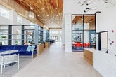 Located at 9295 Ashland Ave in Pensacola, Florida, this single-story community clubhouse showcases a vibrant coastal design aesthetic throughout its expansive interior. The grand entrance opens into a soaring, light-filled lobby adorned with a striking warm wood-planked ceiling, large-format tile flooring, and a sculptural chandelier that sets an impressive tone. A welcoming front desk finished in white stone anchors the entry area, accompanied by casual seating arrangements including wicker chairs and small café tables bathed in natural light from floor-to-ceiling windows. The open lounge area features plush curved sofas, navy blue accent chairs, and an area rug over hardwood flooring, all centered beneath a dramatic installation of hanging rope lights over a communal kitchen island with a herringbone teal tile backsplash. A glass-enclosed conference and co-working room with bold red seating provides a dedicated workspace, while open shelving with navy blue accents lines the main corridor for added functionality. A spacious fitness center is well-appointed with a comprehensive selection of strength training machines, free weights, treadmills, ellipticals, and cardio equipment, all illuminated by clerestory windows and modern linear lighting. An airy café nook features a bold abstract wall mural, coffee station, and ample natural light, completing this thoughtfully designed community hub.