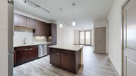 This one-bedroom, one-bathroom unit in San Antonio, TX offers a efficiently designed single-floor layout with a cohesive blend of modern finishes and neutral tones. The open-concept kitchen and living area serves as the heart of the space, anchored by light wood-style plank flooring that extends throughout. The kitchen is well-equipped with rich dark-walnut cabinetry reaching to the ceiling, granite countertops, a glass tile backsplash, stainless steel appliances including a range, microwave, dishwasher, and refrigerator, and a central island with additional counter space. Pendant lighting and under-cabinet lighting add functional illumination. The adjacent living area is generously proportioned, featuring a ceiling fan and track lighting. A hallway connects the main living space to the private bedroom, which features plush carpet flooring, a ceiling fan, and a window offering natural light. The bedroom is served by a spacious walk-in closet outfitted with multiple hanging rods and built-in shelving on all sides. The full bathroom includes a dark-wood vanity with granite countertop, tile flooring, and access to both a soaking tub and a separate tiled shower.
