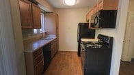 The kitchen is designed with a practical galley-style layout, maximizing efficiency within a compact footprint. Warm-toned wooden cabinetry lines both sides of the space, providing generous upper and lower storage options. The cabinetry features a rich medium-brown finish that adds warmth to the room.

On one side, a double-basin sink sits beneath a small window that allows natural light to filter in, paired with a black dishwasher and granite-look countertops. The opposite side houses a full suite of black appliances, including a coil-burner electric range, an over-the-range microwave, and a full-size refrigerator, all finished in matching black for a cohesive look.

The flooring throughout is a hardwood-style laminate in a warm medium-brown tone, complementing the cabinetry and contributing to an inviting atmosphere. Neutral beige walls keep the space feeling open and bright. Overhead, a flush-mount ceiling light provides even illumination throughout the kitchen. The overall ambiance is functional and well-organized, suited for everyday cooking and meal preparation.