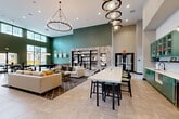 Located at 16200 Croft Drive in Charlotte, NC, this generously sized single-floor community amenity space is designed to impress. Soaring double-height ceilings create an expansive, airy atmosphere throughout, enhanced by abundant natural light streaming in through clerestory windows and large glass doors that open to an outdoor patio area.

The interior is anchored by bold emerald green accent walls that contrast beautifully with neutral tile flooring and warm, contemporary furnishings. A dramatic chandelier and sculptural pendant light fixtures serve as eye-catching focal points, elevating the overall aesthetic.

The space is thoughtfully zoned for multiple uses. A plush lounge area features generous sectional sofas arranged around a textured feature wall with a mounted flat-screen television, ideal for casual gatherings. A billiards table occupies its own defined zone, offering a dedicated recreation area. A long communal high-top table with bar seating provides a functional workspace or social hub, positioned adjacent to a sleek kitchenette outfitted with teal cabinetry, a sink, beverage refrigerator, and pendant lighting. Open shelving with curated décor adds a polished, designer touch throughout. Glass doors lead directly to an outdoor terrace, seamlessly extending the social experience beyond the interior.