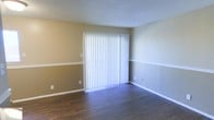 This single-story space presents a clean and functional layout with a neutral, two-tone wall treatment featuring a warm tan lower half and a cooler gray upper half, divided by a crisp white chair rail. The flooring throughout consists of rich, dark hardwood-style planks that add warmth and contrast to the lighter walls. A prominent sliding glass door spans a large portion of one wall, dressed with full-length vertical blinds that allow for adjustable natural light and potential outdoor access. The textured popcorn ceiling adds a classic touch overhead, while white baseboards provide a polished finishing detail along the perimeter. Multiple electrical outlets are distributed throughout the room, and a light switch is conveniently positioned near the doorway. The overall space feels open and versatile, offering a blank canvas suitable for a variety of furniture arrangements. The combination of durable flooring, ample natural light potential, and straightforward design elements creates a practical, welcoming environment that is easy to personalize.