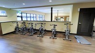This single-floor fitness facility is thoughtfully designed to provide a comprehensive and welcoming workout environment. The cycling studio is a standout feature, equipped with a row of stationary cycling bikes arranged on smooth hardwood-style flooring, creating an organized and energizing space for group cycling sessions. Large windows along the studio walls invite generous amounts of natural light, contributing to an open and motivating atmosphere. The studio is finished with neutral-toned walls and recessed lighting overhead, ensuring a bright and comfortable environment at any time of day. A low partition wall separates the cycling area from an adjacent space, which appears to include additional fitness equipment and a mirrored wall — a common feature that enhances the sense of space and supports form correction during workouts. Dark floor trim and structural columns add clean, architectural contrast throughout the interior. The facility's layout promotes an efficient flow between workout zones, making it easy for users to transition between activities. Overall, the space reflects a practical yet polished design, well-suited for both individual training and group fitness programming.