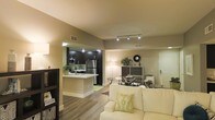 This single-floor residence offers a thoughtfully designed open-concept layout that effortlessly connects the kitchen, dining, and living areas into one cohesive and functional space. The living area is anchored by a plush seating arrangement and benefits from warm ambient lighting that creates a welcoming atmosphere throughout. Rich hardwood-style flooring runs continuously across the space, adding warmth and visual continuity.

The kitchen is a standout feature, showcasing dark espresso cabinetry paired with stainless steel appliances, including a full-size refrigerator and microwave. A peninsula counter provides additional prep space and serves as a natural divide between the kitchen and the rest of the living area. Track lighting overhead ensures the kitchen is well-lit and functional.

Adjacent to the living space, a glass-top dining table with coordinating chairs creates an intimate area for meals and gatherings. Decorative accents on a nearby console table add personality and a sense of style to the space. Neutral-toned walls throughout enhance the brightness and openness of the interior, while multiple light sources contribute to a warm and inviting ambiance. The layout is efficient yet spacious, making excellent use of the available footprint.