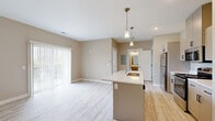 Located at 3800 E Campus Ave in Holland, Michigan, this two-bedroom, one-bathroom residence offers a thoughtfully designed single-floor layout with a clean, modern aesthetic. The open-concept living and kitchen area forms the heart of the space, anchored by a central kitchen island with a white quartz countertop and an undermount sink, illuminated by elegant pendant lighting with brass accents. The kitchen is well-equipped with stainless steel appliances — including a refrigerator, gas range, and microwave — complemented by taupe cabinetry and light wood-look flooring that extends throughout the main living area. A sliding glass door with vertical blinds brings in natural light and connects the living space to the outdoors. A dedicated in-unit laundry closet with a stacked washer and dryer adds everyday convenience. Both bedrooms feature neutral carpet flooring, warm beige walls, and windows with blinds that allow for natural light and privacy. The primary bedroom includes a spacious walk-in closet with built-in wire shelving. The bathroom is finished with light wood-look tile flooring, a gray vanity with a white countertop, a framed mirror, and a shower-tub combination.