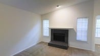 This single-story space presents a straightforward, functional layout with neutral design choices throughout. The main living area is anchored by a dark brick fireplace, which serves as a natural focal point. The fireplace features a raised brick hearth and a simple white mantel shelf, adding a classic structural element to the room. The walls are finished in a clean, light neutral tone, complementing the wall-to-wall carpeting in a matching beige palette, creating a cohesive and understated aesthetic. The textured ceiling adds subtle dimension to the space. Multiple windows fitted with white horizontal blinds allow natural light to filter into the room, contributing to an open and airy feel. The vaulted ceiling height above the fireplace wall adds a sense of vertical space, making the living area feel more expansive. The overall design is minimal and move-in ready, providing a blank canvas well-suited for a variety of interior styles. The combination of the brick fireplace, neutral tones, and carpeted flooring creates a comfortable, livable environment throughout this single-floor space.