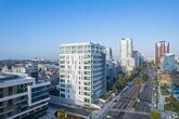 Located at 200 W Ocean Blvd in Long Beach, California, this contemporary fitness studio offers a bright and energizing environment designed for a well-rounded workout experience. The space is defined by dramatic floor-to-ceiling glass walls on multiple sides, flooding the interior with abundant natural light and providing open views of the surrounding urban landscape, including palm trees and neighboring architecture — a fitting backdrop for an active lifestyle near the Southern California coast.

The light hardwood flooring runs throughout, lending warmth and a clean aesthetic to the room. Above, a striking ceiling treatment features parallel dark acoustic baffles set against a white structure, enhancing both the visual design and the acoustic quality of the space.

The studio is equipped with a thoughtful selection of fitness equipment, including rowing machines, stationary spin bikes, and a multi-function power rack, catering to both cardio and strength training needs. A dedicated accessory zone features open shelving stocked with medicine balls, resistance tools, and wellness accessories, alongside stability balls and a dumbbell rack. A wall-mounted mirror adds functionality and amplifies the sense of openness. The glass-paneled entry door maintains visual connectivity to adjacent spaces while keeping the studio feeling light and airy throughout.