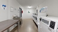 The laundry room at 8 Wilsonview Ave is a well-organized, single-floor shared facility designed for efficient everyday use. The space is laid out with a clear, functional flow, beginning with a corridor entry that leads into the main laundry area. The room features a row of modern front-loading washing machines and dryers, all finished in white, neatly aligned along one wall and equipped with card or coin-operated controls. Overhead fluorescent tube lighting provides bright, even illumination throughout the space. The floors are finished with durable speckled linoleum, well-suited to the demands of a high-traffic utility area. A stainless steel utility sink and a wooden folding table are positioned along the opposite wall, offering practical workspaces for sorting and handling laundry. Exposed plumbing and ventilation ductwork are visible along the back wall behind the machines. Instructional and informational notices are posted throughout, including water-saving guidelines and machine usage instructions. A fire safety pull station is mounted prominently near the maintenance room door, which is clearly labeled and accessible from within the space. The white walls and clean layout contribute to a tidy, no-frills environment that prioritizes practicality and ease of use.