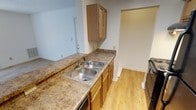 This single-floor space showcases a well-appointed kitchen as a central feature. The kitchen is equipped with warm-toned wooden cabinetry, both upper and lower, providing ample storage for everyday essentials. The countertops display a granite-style surface with earthy brown and gold tones, offering generous prep space that extends along the length of the kitchen. A double-basin stainless steel sink sits along the counter, paired with a standard faucet, and is conveniently positioned beneath an upper cabinet. The kitchen also accommodates standard appliances, including what appears to be a dishwasher and additional appliance to the right. Light hardwood flooring runs throughout the space, contributing a clean and cohesive aesthetic. The walls are finished in a neutral light gray tone, enhancing the bright and open feel of the interior. An open passageway visible from the kitchen leads to an adjacent area, suggesting a practical, connected layout. A door with a light-colored frame is visible in the background, indicating access to another room or exterior. Overall, the space presents a straightforward, functional design with consistent finishes throughout.