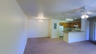 This single-floor space offers an open layout that seamlessly connects the living area to the kitchen. The living area is finished with plush carpeting in a neutral mauve tone, creating a warm and comfortable atmosphere. A small chandelier serves as a subtle decorative focal point overhead, adding a touch of character to the otherwise clean, white-walled space.

The kitchen is positioned toward the rear and is defined by a peninsular counter that provides both workspace and a natural visual divider between the two areas. Warm, honey-toned wooden cabinetry lines the upper walls, complementing the light countertops. A white refrigerator and a built-in cooktop are among the visible appliances, with a ventilation hood mounted above the range area. Flush fluorescent lighting illuminates the kitchen workspace effectively.

An electrical panel is accessible along one wall, and a visible ceiling vent suggests a centralized HVAC system. The overall aesthetic is functional and straightforward, with white walls and ceilings throughout that lend brightness to the interior. The open flow between the living and kitchen areas makes the space feel practical and easy to navigate.
