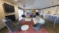 This contemporary community lounge space is designed to accommodate a variety of activities, seamlessly blending work-ready amenities with comfortable social areas. At the forefront is a large, light-wood conference table fitted with built-in power access points and surrounded by modern grey chairs, making it ideal for meetings or collaborative work sessions. Beyond the table, a sleek beverage station — equipped with coffee urns and a bar-height counter with stools — serves as a central hub for refreshments. The lounge area features a generously sized white sofa adorned with accent pillows, flanked by contemporary grey armchairs, all arranged atop a bold, patterned red area rug. Two distinctive sputnik-style pendant light fixtures hang overhead, casting a warm, stylish glow throughout the open-plan interior. Vaulted ceilings and large windows enhance the sense of openness and allow natural light to filter in. A staircase with dark treads adds architectural interest to the background, while tasteful decorative elements — including wall art and sculptural accents — contribute to the space's modern, curated aesthetic. The dark hardwood flooring grounds the space with a polished, cohesive finish throughout.