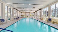 This indoor pool facility offers a well-appointed aquatic environment designed for year-round use. The centerpiece is a generously sized rectangular lap pool with clear, turquoise water, surrounded by wide tiled decking that provides ample space for movement and relaxation. A standout architectural feature is the row of full-height retractable glass garage doors lining both sides of the pool, allowing the space to open up to the outdoors when weather permits, flooding the interior with natural light and views of the surrounding community buildings. When closed, the expansive glazing maintains a bright, airy atmosphere throughout.

The ceiling is finished with white paneling and fitted with linear fluorescent lighting, ensuring consistent illumination across the entire pool area. Along the poolside decking, blue sling-style lounge chairs and small round side tables are arranged for convenient poolside relaxation. A dedicated lounge area at one end of the facility features additional reclining chairs and paired seating arrangements. Safety signage, an emergency cutoff switch, a water fountain, and a double-door entry are also present. Pool cleaning equipment is stored on-site, and safety buoys are mounted on the walls, reflecting a well-maintained and safety-conscious facility.