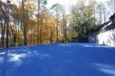 This outdoor recreational facility offers a well-appointed multi-sport court designed to accommodate a variety of athletic activities. The expansive blue-surfaced playing area is clearly marked with court lines suitable for tennis and other court sports, while a portable soccer goal positioned at one end enables small-sided soccer play as well. The court is fully enclosed by a sturdy chain-link fence, providing a safe and defined boundary for all activities. Tall trees surround the perimeter, offering natural shade and a pleasant, park-like ambiance that softens the urban setting. Along one side, a textured stucco retaining wall runs the length of the court, topped by a raised walkway with decorative black metal railings and a pergola structure, adding architectural character to the space. Overhead court lighting poles ensure the facility can be enjoyed during evening hours. A neighboring red-brick building and mature landscaping are visible beyond the fencing, situating the court within a residential community setting. The space reflects a commitment to outdoor recreation and community engagement, making it a practical and inviting destination for sports enthusiasts of all ages.