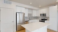 This thoughtfully designed single-floor unit offers a clean, modern aesthetic across its well-utilized layout. The open-concept kitchen and living area serves as the heart of the space, anchored by a central island with a sleek quartz countertop — ideal for both meal prep and casual dining. The kitchen is fully equipped with stainless steel appliances, including a French door refrigerator, electric range, built-in microwave, and dishwasher. Crisp white shaker-style cabinetry lines the walls, complemented by a light gray subway tile backsplash that adds subtle texture. Recessed lighting illuminates the space efficiently throughout. The living area benefits from large windows and a sliding glass door that opens to a balcony, flooding the room with natural light and offering views of the surrounding neighborhood. Light-toned hardwood-style flooring runs consistently throughout the entire unit, creating a cohesive and airy feel. The bedroom is a comfortable size, featuring a ceiling fan, a window with exterior views, and direct access to a dedicated walk-in closet equipped with built-in shelving and hanging rods. A bathroom with vanity cabinetry is conveniently accessible from the bedroom area. The overall palette — white walls, light floors, and neutral tones — gives the space a bright, contemporary character.