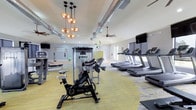 This modern fitness center is thoughtfully designed to accommodate a wide range of workout styles and fitness goals. The space is organized into distinct functional zones, each outfitted with quality equipment and finished with stylish, durable materials throughout.

The cardio area features a row of treadmills positioned along large windows that bring in natural light and offer views of the surrounding greenery, keeping the space feeling open and energized. Wall-mounted flat-screen televisions are stationed above the treadmills for entertainment during workouts. Ceiling fans provide additional air circulation throughout.

Adjacent to the cardio zone, a dedicated strength training area houses multiple cable and resistance machines, including Torque functional training systems, along with a free weights section complete with a dumbbell rack and adjustable bench. The flooring transitions between patterned carpet tiles in grey and yellow tones and smooth hardwood, clearly delineating the different workout areas.

A functional training and flexibility zone features suspension training straps mounted to the wall, a stability ball, and an open hardwood floor space ideal for stretching, yoga, or bodyweight exercises. Decorative accent walls in chartreuse green add a vibrant, energetic touch to the interior aesthetic.

Industrial-style exposed ductwork runs along the ceilings, complemented by pendant cluster lighting fixtures that add a contemporary design element. The overall ambiance is bright, clean, and motivating.