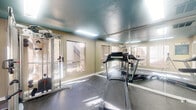 The Flint & 290 Fitness Center, located at 5915 Flintlock Rd in Houston, TX, is a dedicated workout facility designed to provide residents with a functional and well-equipped exercise space. The room is contained within a single-level layout and makes efficient use of its compact footprint.

The interior features a distinctive teal-painted textured ceiling, which adds a pop of color and visual contrast against the neutral, light wood-paneled walls. Durable, dark rubber flooring covers the entire space, offering both safety and practicality for workout activities. Flush-mounted fluorescent light panels illuminate the room evenly, supplemented by abundant natural light streaming in through multiple large windows that line the walls and offer views of the surrounding property.

A defining architectural feature is the full floor-to-ceiling mirrored wall panel, which visually expands the space and allows users to monitor their form during exercise. The facility includes cardio equipment such as treadmills, as well as a multi-station cable weight machine for strength training. A wall-mounted split air conditioning unit ensures climate-controlled comfort year-round.

A glass entry door provides easy access, and posted fitness center rules ensure a shared, respectful environment. The space is bright, practical, and thoughtfully arranged to accommodate a variety of fitness routines within a streamlined, community-oriented setting.