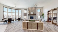 This single-floor community clubhouse blends modern comfort with warm architectural details throughout its well-appointed spaces. The centerpiece of the main lounge is a dramatic floor-to-ceiling stacked stone fireplace with a wood mantel, flanked by seating areas beneath dual ceiling fans and recessed lighting. A sweeping curved wall of wood-trimmed bay windows floods the dining area with abundant natural light and offers panoramic views of the surrounding neighborhood.

The kitchen is a functional showpiece, featuring warm two-tone cabinetry, a large white quartz island with an undermount sink and bar seating, stainless steel appliances, and striking sputnik pendant lighting that reaches toward the soaring vaulted ceiling. Tile flooring grounds the space with a clean, polished aesthetic. An adjacent conference or meeting room offers a round table, comfortable seating, and vibrant wall art, making it ideal for gatherings or private meetings.

The game and media rooms provide relaxed entertainment options, with plush carpeting, wainscoting panels, wood-trimmed windows, and a wall-mounted television. A covered outdoor patio with heavy timber framing and a tongue-and-groove ceiling offers a welcoming entrance and additional seating, completing this well-rounded communal facility.