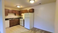 This single-floor space includes a kitchen designed for practical everyday use. The kitchen is outfitted with rich, warm-toned wood cabinetry that provides generous upper and lower storage, complemented by dark countertops that offer a subtle contrast. A double-basin sink is positioned along the lower cabinetry, while the layout follows an efficient L-shaped configuration. White appliances are included, featuring an electric range with overhead ventilation, and a full-sized refrigerator that anchors one end of the kitchen. Flush-mount ceiling light fixtures provide consistent overhead illumination throughout the space. The flooring in the kitchen area consists of tile in a neutral tone, transitioning to carpeting in the adjacent area, clearly delineating the functional zones within the unit. The walls are painted in a clean, neutral off-white, contributing to a bright and open atmosphere. The overall design of the space is straightforward and utilitarian, prioritizing function and ease of use. Ceiling-mounted HVAC vents suggest a central climate control system, adding to the practical nature of this well-maintained space.