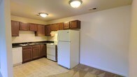 This single-floor space includes a kitchen designed for practical everyday use. The kitchen is outfitted with rich, warm-toned wood cabinetry that provides generous upper and lower storage, complemented by dark countertops that offer a subtle contrast. A double-basin sink is positioned along the lower cabinetry, while the layout follows an efficient L-shaped configuration. White appliances are included, featuring an electric range with overhead ventilation, and a full-sized refrigerator that anchors one end of the kitchen. Flush-mount ceiling light fixtures provide consistent overhead illumination throughout the space. The flooring in the kitchen area consists of tile in a neutral tone, transitioning to carpeting in the adjacent area, clearly delineating the functional zones within the unit. The walls are painted in a clean, neutral off-white, contributing to a bright and open atmosphere. The overall design of the space is straightforward and utilitarian, prioritizing function and ease of use. Ceiling-mounted HVAC vents suggest a central climate control system, adding to the practical nature of this well-maintained space.
