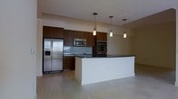 This single-floor space showcases a thoughtfully designed open-concept layout centered around a stunning modern kitchen. Rich, dark wood cabinetry lines the walls, providing ample storage while lending a warm, sophisticated contrast to the light-toned hardwood flooring that flows throughout the space. The expansive kitchen island features a crisp white base topped with a sleek dark countertop, offering generous workspace and a natural gathering point. Three elegant pendant lights hang in a row above the island, casting a warm, focused glow that enhances the kitchen's contemporary aesthetic. The kitchen is equipped with stainless steel appliances, including a French door refrigerator with an in-door dispenser, a built-in microwave, and a double wall oven, all seamlessly integrated into the cabinetry. A glass tile backsplash adds a refined touch behind the cooktop area. The neutral wall tones and light flooring create an airy, open ambiance, while the clean lines and minimalist design give the space a polished, modern character. The overall layout promotes a seamless flow, making the kitchen both highly functional and visually impressive.