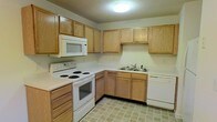 This single-floor space includes a kitchen designed with functionality in mind. The kitchen features an L-shaped layout that makes efficient use of the available space, providing a logical workflow between the cooking, preparation, and cleaning zones. Light oak wood cabinetry lines the upper and lower walls, offering generous storage throughout. White appliances, including an electric coil range, an over-the-range microwave, and a dishwasher, are seamlessly integrated into the design. The double-basin sink is positioned along the lower cabinetry run, complemented by a standard faucet. Countertops in a clean, light finish run continuously along the layout, providing ample prep space. The flooring consists of neutral-toned tile in a grid pattern, contributing to the kitchen's understated and practical aesthetic. A textured ceiling with a flush-mounted light fixture provides even overhead illumination throughout the space. The overall ambiance of the kitchen is straightforward and utilitarian, offering a clean and organized environment well-suited for everyday use.
