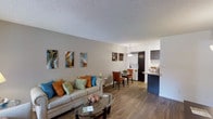 This single-floor space showcases an open-concept layout that connects the living room, dining area, and kitchen in one continuous, flowing environment. Hardwood-style flooring runs throughout, lending warmth and visual cohesion to the entire space.

The living area is comfortably sized, anchored by a neutral-toned sofa adorned with vibrant teal, orange, and green accent pillows that add pops of color. A round glass coffee table sits at the center, and a curated arrangement of abstract wall art adds personality to the light gray walls. The textured ceiling adds subtle architectural detail overhead.

Transitioning seamlessly into the dining area, a compact table with burnt-orange upholstered chairs sits beneath a pendant light fixture, creating an intimate setting for meals. Decorative wall art echoes the colorful theme carried throughout the space.

The kitchen is visibly integrated into the open floor plan, featuring dark cabinetry, a stainless steel refrigerator, and a peninsula counter with a navy blue base, providing a natural divider while maintaining the open feel. The overall ambiance is modern, warm, and inviting, with a cohesive color palette that ties each zone together effortlessly.