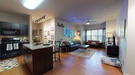 Located at 80 E Exchange St in downtown Akron, Ohio, this single-floor unit offers a well-designed open-concept layout that seamlessly connects the kitchen, dining, and living areas. The living room is a welcoming space featuring hardwood-style flooring, a ceiling fan, large windows that allow ample natural light, and stylish décor including a bold neon accent piece. Two sofas, a coffee table, and a vibrant area rug complete the comfortable gathering space.

The kitchen is outfitted with dark espresso cabinetry, granite-style countertops, black appliances including a range, built-in microwave, and dishwasher, all set atop hardwood-style flooring. A kitchen island with seating provides additional prep and dining space.

Multiple bedrooms offer plush carpeting, natural light through large windows with blinds and curtains, and ample room for full or queen-sized beds with accompanying furniture. Each room provides a cozy, private retreat.

Two full bathrooms feature tub-shower combinations, hardwood-style flooring, dark vanity cabinetry with granite-style countertops, and tasteful decorative accents. The cohesive design throughout the unit balances modern finishes with a warm, livable atmosphere.