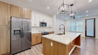 Located at 2031 Synergy Lane in Sarasota, FL, this thoughtfully designed one-bedroom, one-bathroom residence offers a modern aesthetic with high-quality finishes throughout its single-floor layout. The kitchen serves as the centerpiece of the home, showcasing warm natural wood cabinetry paired with crisp white upper cabinets, a striking veined quartz backsplash and countertops, and a large central island with an undermount sink and matte black faucet. Stainless steel appliances, including a French door refrigerator, range, and built-in microwave, complete the culinary space, while geometric black iron pendant lights add an industrial-chic touch overhead. Light wood-look flooring flows throughout the main living areas, complementing the clean white walls and recessed lighting. The living area features a ceiling fan and a bright window with blinds, allowing ample natural light. The bedroom is carpeted for comfort, offering a ceiling fan, generous natural light from a large window, and a dedicated closet. The bathroom is well-appointed with white shaker cabinetry, quartz countertops, matte black hardware, wood-look tile flooring, and a subway-tiled soaking tub and shower combination. A dedicated laundry area rounds out this efficiently designed, move-in-ready home.