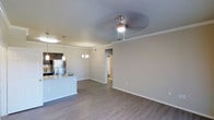 This single-floor residence showcases an open-concept layout that connects the living area and kitchen in one cohesive, flowing space. The living area is finished with warm hardwood-style flooring that extends throughout, complemented by crisp white baseboards and crown molding that add a refined architectural detail. A ceiling fan with an integrated light fixture anchors the living space, while a decorative chandelier adds an elegant touch to the dining area nearby.

The kitchen is positioned to the left, featuring dark cabinetry, a stainless steel refrigerator, and a peninsula with granite-style countertops and pendant lighting above — ideal for casual dining or meal preparation. The neutral, light-toned walls create a bright and welcoming ambiance throughout, enhancing the sense of openness.

A hallway visible from the main living area leads to additional rooms, suggesting a thoughtful and functional floor plan. The overall design balances contemporary finishes with practical layout choices, making the space feel both stylish and livable.
