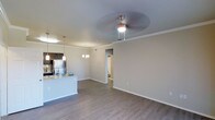This single-floor residence showcases an open-concept layout that connects the living area and kitchen in one cohesive, flowing space. The living area is finished with warm hardwood-style flooring that extends throughout, complemented by crisp white baseboards and crown molding that add a refined architectural detail. A ceiling fan with an integrated light fixture anchors the living space, while a decorative chandelier adds an elegant touch to the dining area nearby.

The kitchen is positioned to the left, featuring dark cabinetry, a stainless steel refrigerator, and a peninsula with granite-style countertops and pendant lighting above — ideal for casual dining or meal preparation. The neutral, light-toned walls create a bright and welcoming ambiance throughout, enhancing the sense of openness.

A hallway visible from the main living area leads to additional rooms, suggesting a thoughtful and functional floor plan. The overall design balances contemporary finishes with practical layout choices, making the space feel both stylish and livable.