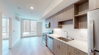 This contemporary single-floor unit is thoughtfully designed with a clean, modern aesthetic throughout. The open-plan kitchen flows seamlessly into the living area, creating a cohesive and functional living space. The kitchen is outfitted with sleek, wood-toned cabinetry that extends to the ceiling for ample storage, complemented by open shelving, white quartz countertops, and a full suite of stainless steel appliances including a range, microwave, dishwasher, and refrigerator. Light hardwood flooring runs throughout the unit, adding warmth and continuity to the space.

The living area is anchored by a large floor-to-ceiling window that floods the room with natural light and offers views of the surrounding urban streetscape. A wall-mounted split air conditioning unit ensures year-round comfort. Adjacent to the kitchen, a dedicated walk-in closet with built-in wire shelving provides generous storage solutions.

The full bathroom is finished with the same attention to detail found throughout the unit, featuring a wood-toned vanity with a white quartz countertop, an undermount sink with modern fixtures, a large mirror, and a full tub-shower combination. Clean white walls and wood-look tile flooring complete the space, maintaining a cohesive and polished look from room to room.