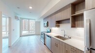 This contemporary single-floor unit is thoughtfully designed with a clean, modern aesthetic throughout. The open-plan kitchen flows seamlessly into the living area, creating a cohesive and functional living space. The kitchen is outfitted with sleek, wood-toned cabinetry that extends to the ceiling for ample storage, complemented by open shelving, white quartz countertops, and a full suite of stainless steel appliances including a range, microwave, dishwasher, and refrigerator. Light hardwood flooring runs throughout the unit, adding warmth and continuity to the space.

The living area is anchored by a large floor-to-ceiling window that floods the room with natural light and offers views of the surrounding urban streetscape. A wall-mounted split air conditioning unit ensures year-round comfort. Adjacent to the kitchen, a dedicated walk-in closet with built-in wire shelving provides generous storage solutions.

The full bathroom is finished with the same attention to detail found throughout the unit, featuring a wood-toned vanity with a white quartz countertop, an undermount sink with modern fixtures, a large mirror, and a full tub-shower combination. Clean white walls and wood-look tile flooring complete the space, maintaining a cohesive and polished look from room to room.
